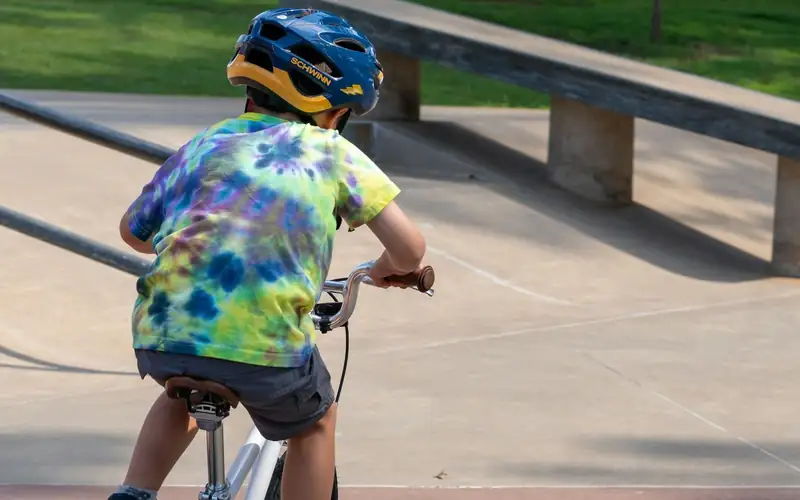 Kind met blauwe helm en tie-dye shirt fietst over een betonpad, van achteren gezien