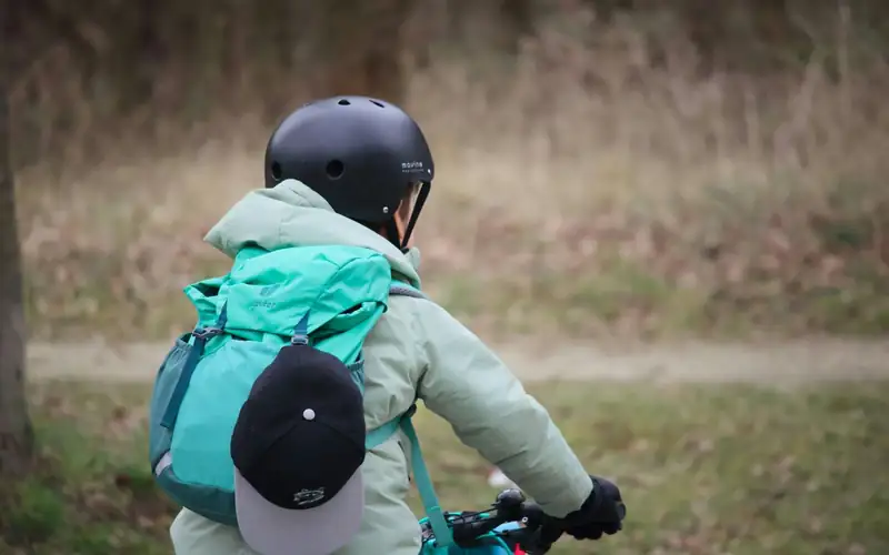 Kind met donkere helm en mintgroene rugzak op de fiets, van achteren gezien