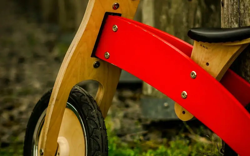 Houten loopfiets met rood frame en zwarte handvatten tegen een schutting
