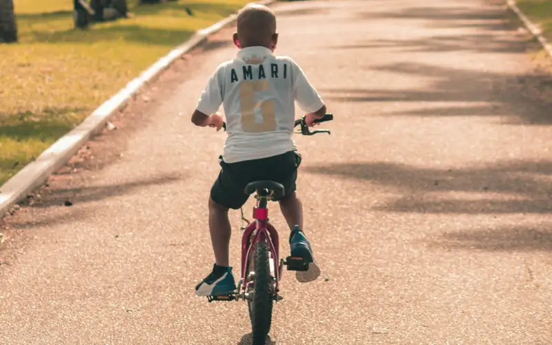 Kind met helm fietst op een kleine rode fiets over een breed parkpad, van achteren gezien