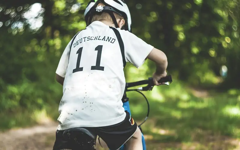 Kind met witte helm en voetbalshirt fietst op een blauwe mountainbike door het bos, van achteren gezien