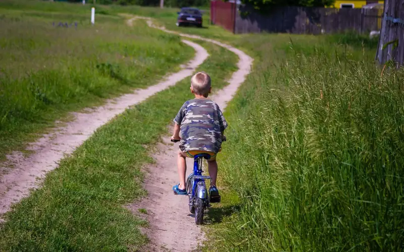 Kind fietst van de camera weg over een zandpad tussen hoog gras
